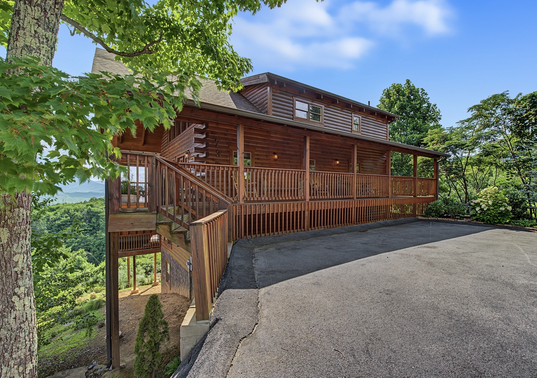 A two-story cabin nestled among the trees features a spacious wrap-around deck and paved parking, with mountain views stretching across the horizon.