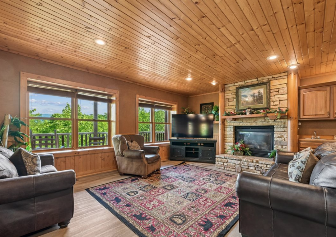 Sink into plush leather sofas before a warm stone fireplace, with natural light streaming through large windows overlooking lush greenery. Your cozy cabin retreat with modern comforts awaits.