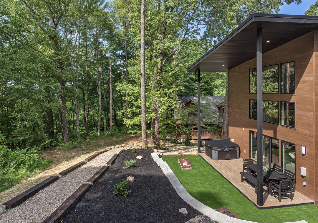 The modern vacation home sits nestled among towering trees, featuring a spacious backyard with manicured lawn, landscaped garden beds, and inviting hot tub on the deck—perfect for relaxing after exploring the surrounding forest.