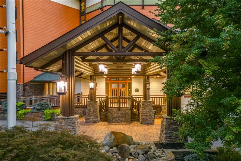 Welcoming covered entrance with timber beams, stone columns, and warm lighting creates an inviting arrival experience surrounded by lush landscaping.