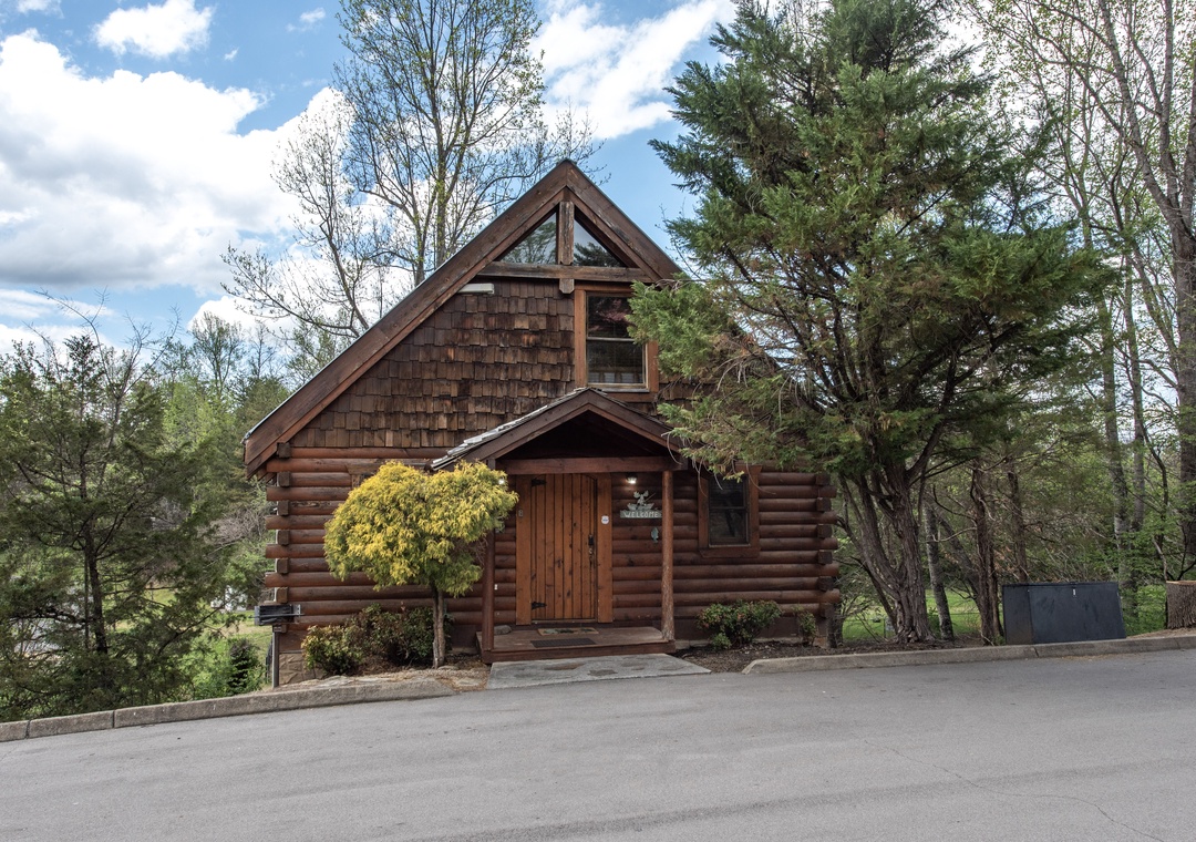 A charming log cabin retreat nestled among towering trees, featuring rustic wood siding and convenient parking for a perfect mountain getaway.