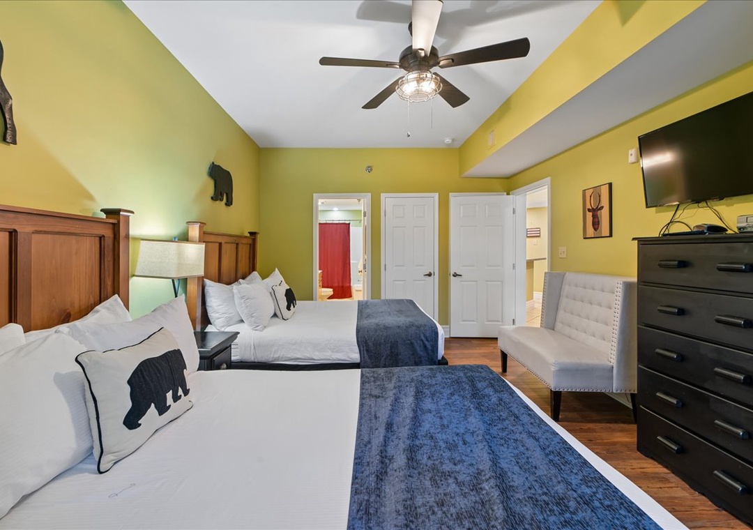 Your cozy retreat features two comfortable beds with rustic wood headboards, cheerful yellow walls, and modern amenities like a flat-screen TV and ceiling fan for your comfort.