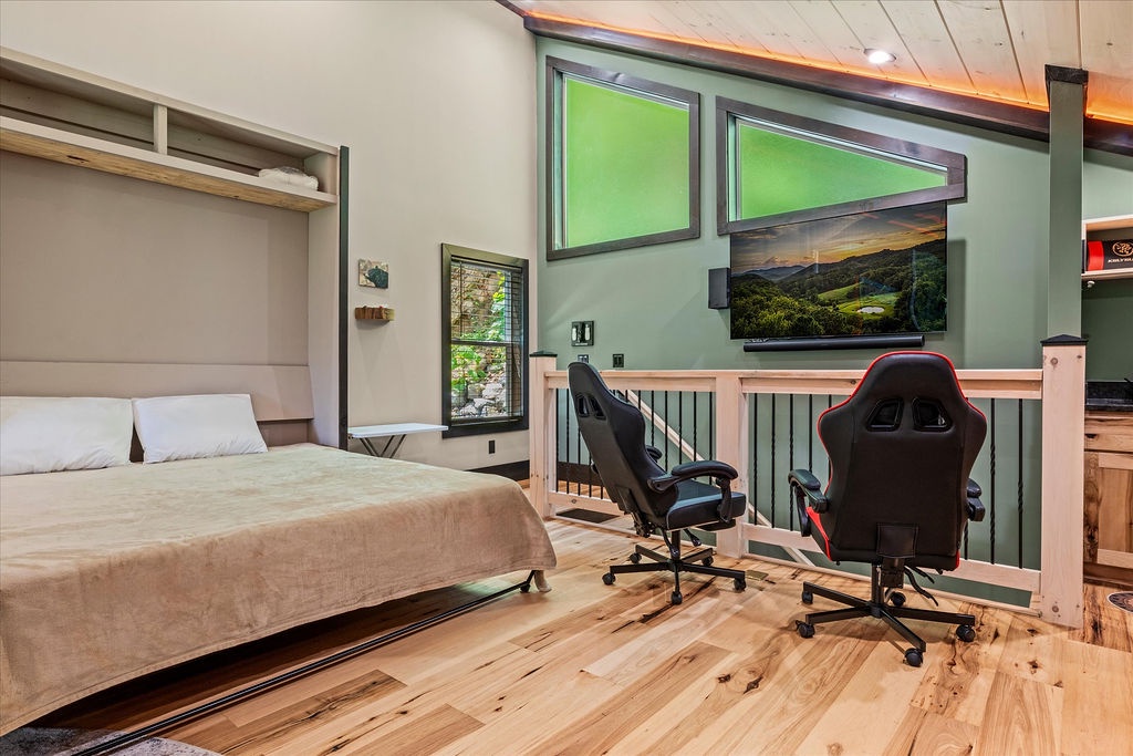 Step into your loft bedroom where a cozy murphy bed meets gaming-ready racing chairs—a unique space with warm wood floors, vaulted ceilings, and a big-screen TV for unwinding after adventures.