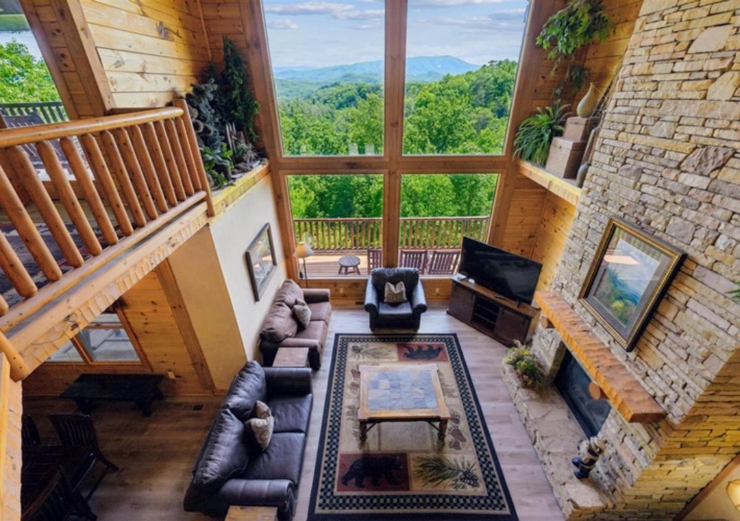 The stunning mountain vistas framed by floor-to-ceiling windows create a serene backdrop for this spacious cabin living area, where warm wood tones and a stone fireplace invite relaxation after your day of exploring.