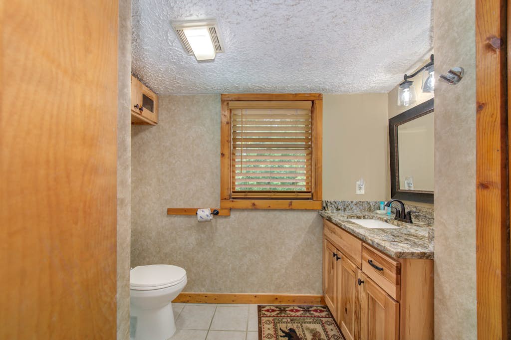 Your private bathroom features warm wood cabinetry, granite countertops, natural light from a charming wood-framed window, and a clean, cozy atmosphere that makes freshening up feel like home.