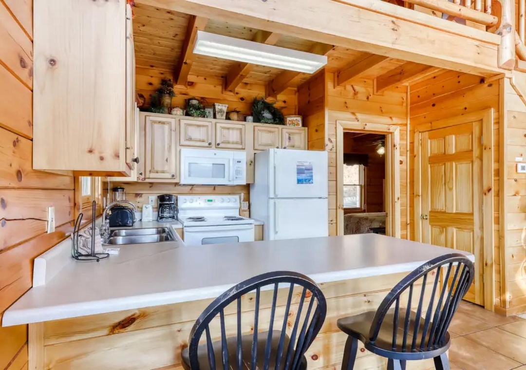 Cozy cabin kitchen where rustic charm meets modern convenience. Warm wood beams frame the space, with a full kitchen, breakfast bar, and inviting gathering spot for meals and morning coffee.
