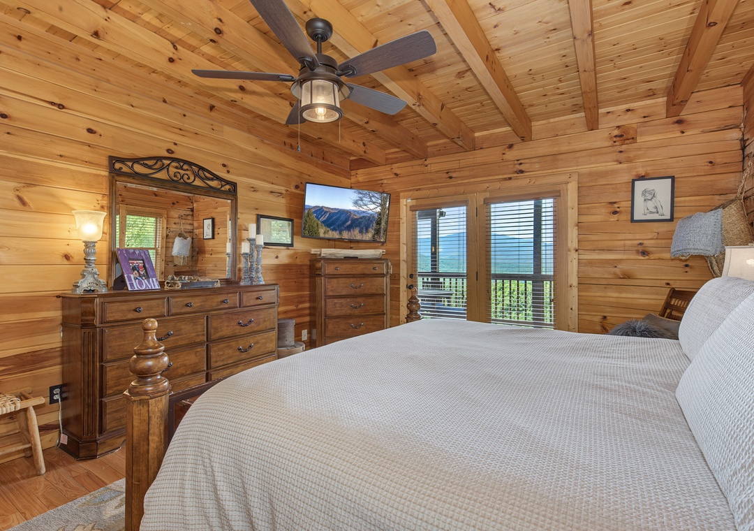 Wake up in your cozy cabin bedroom with knotty pine walls, a ceiling fan above, and mountain views framed by your private balcony doors. The rustic charm and fresh air make mornings feel magical.