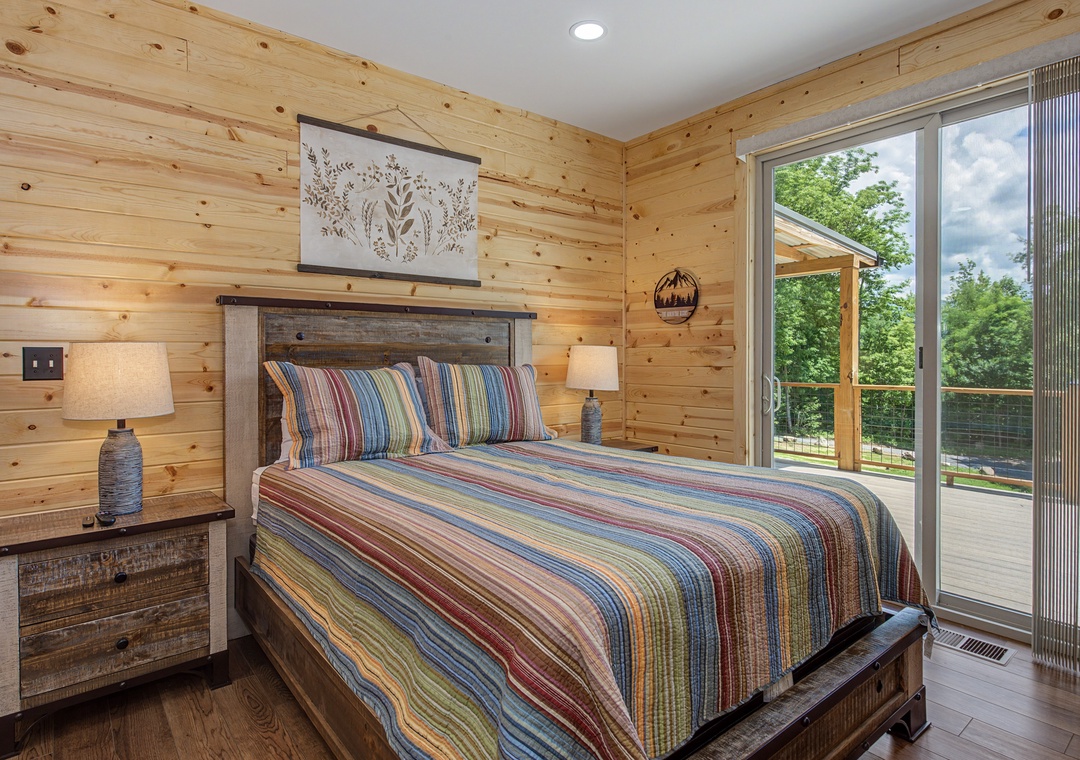 Wake up to natural light streaming through your private balcony doors, where colorful linens and warm wood walls create a cozy retreat with peaceful woodland views just beyond.