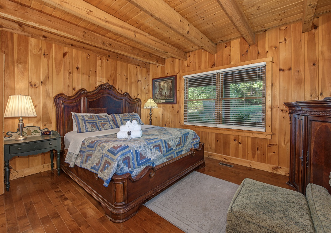 Your cozy bedroom retreat features warm wood-paneled walls, vaulted ceilings, and a grand carved bed dressed in handmade quilts. Natural light streams through the window, while soft lamplight invites you to unwind in this peaceful cabin sanctuary.