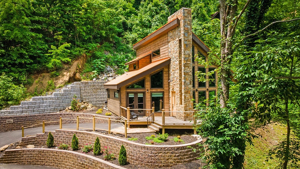 A modern cabin nestled in lush forest, featuring warm wood siding, stone chimney, and terraced landscaping that blends seamlessly with natural surroundings.
