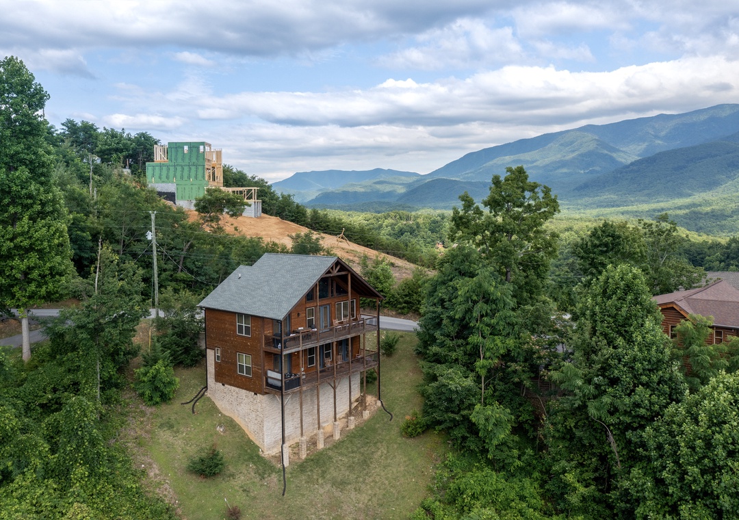 Aerial view of a charming mountain cabin nestled among lush trees with sweeping views of forested peaks and valleys in the distance.