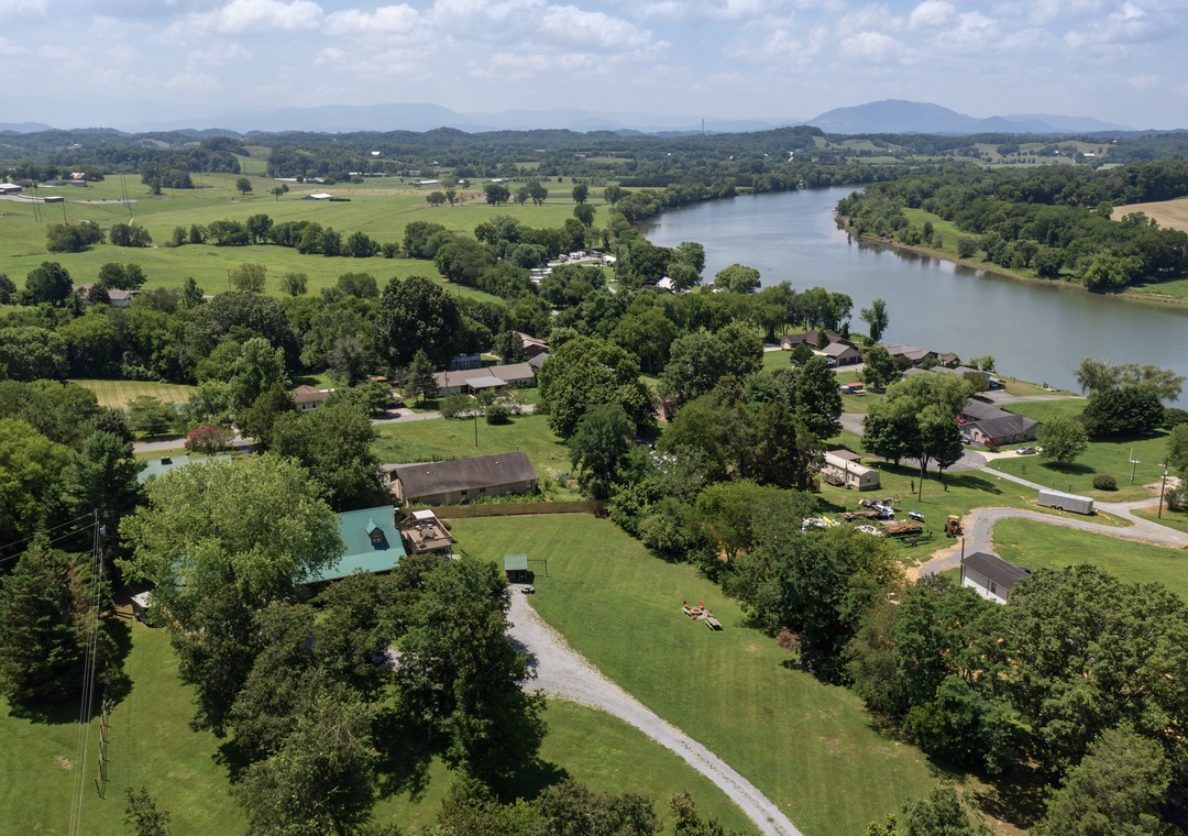 Aerial view of a peaceful riverside neighborhood surrounded by rolling farmland, lush forests, and distant mountain ridges—a tranquil rural setting perfect for escaping the everyday.
