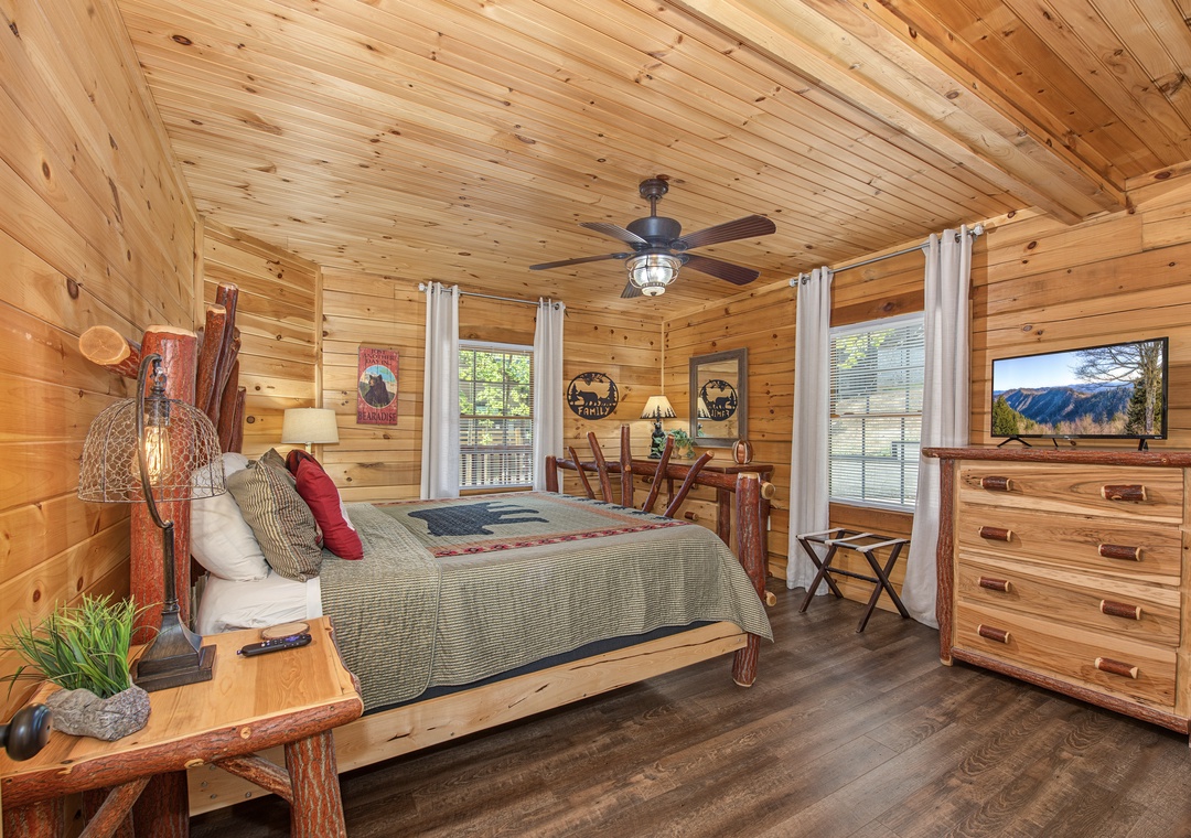 Unwind in your cozy cabin bedroom with warm wood walls, rustic furnishings, and natural light streaming through large windows. Ceiling fan keeps you comfortable year-round.
