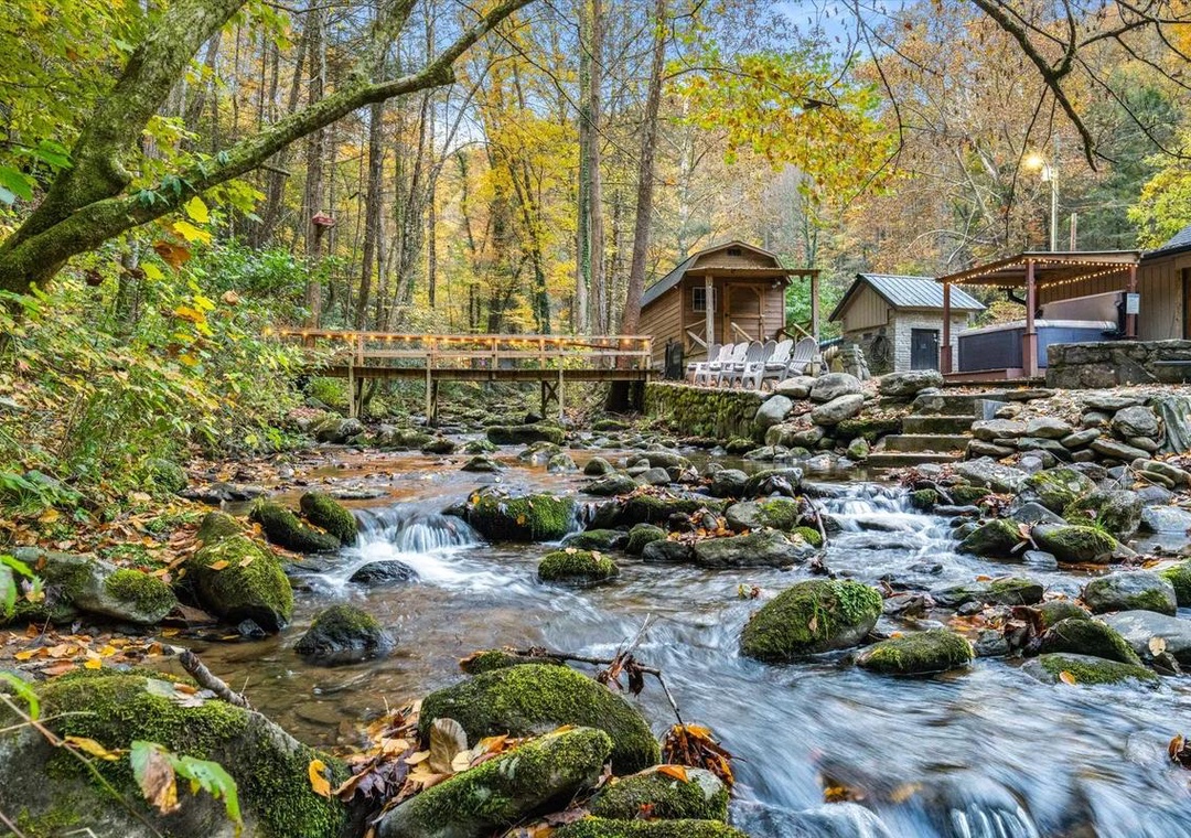 A serene creek flows through the property's wooded grounds, passing beneath a rustic footbridge with autumn foliage framing the peaceful setting.