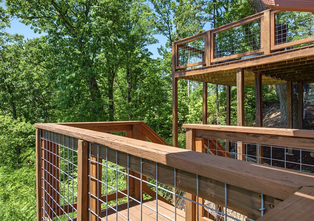 Multi-level wooden deck surrounded by lush forest trees. The deck features wire mesh railing and natural wood construction, positioned against a wooded hillside backdrop.