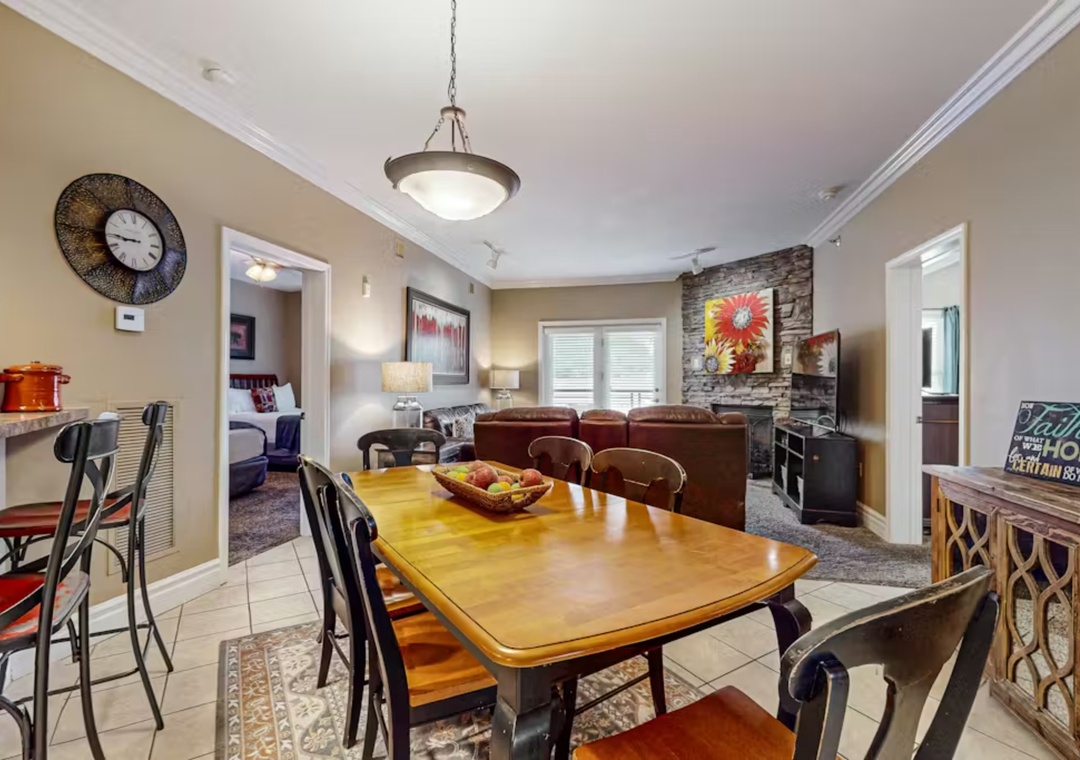 Gather around the warm wooden dining table where your meals become memories, with cozy living space and stone fireplace creating the perfect home atmosphere.