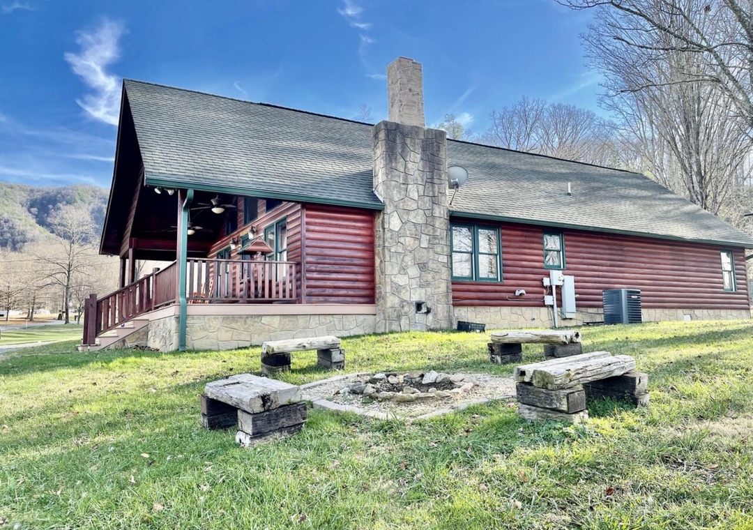 A charming log cabin sits nestled in scenic surroundings, featuring a covered porch, outdoor fire pit with rustic seating, and stunning mountain views that frame the property's peaceful setting.