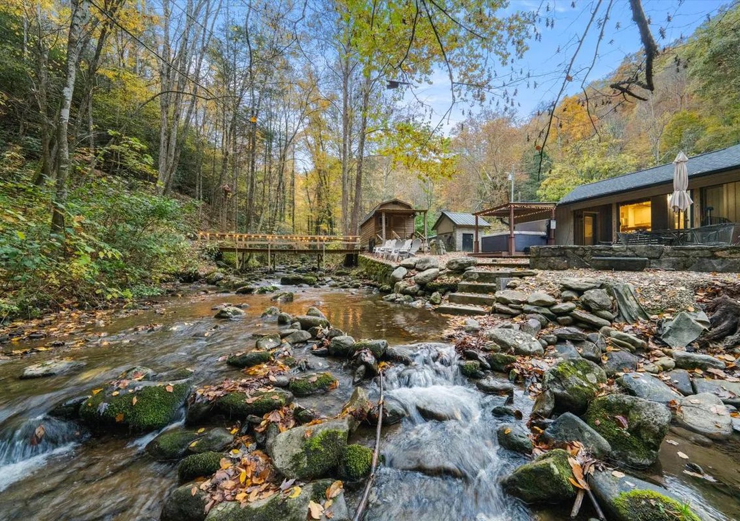 A serene mountain stream flows through the property grounds, surrounded by autumn foliage and natural stone formations, with a charming footbridge connecting wooded trails to cozy guest accommodations.