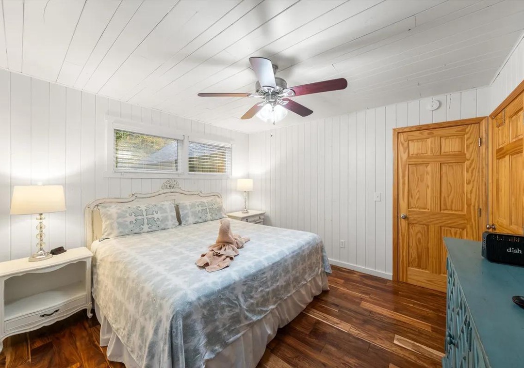 Serene bedroom retreat with soft white paneled walls, rich hardwood floors, and natural light. Vintage-style furnishings and a ceiling fan create a comfortable, welcoming space.