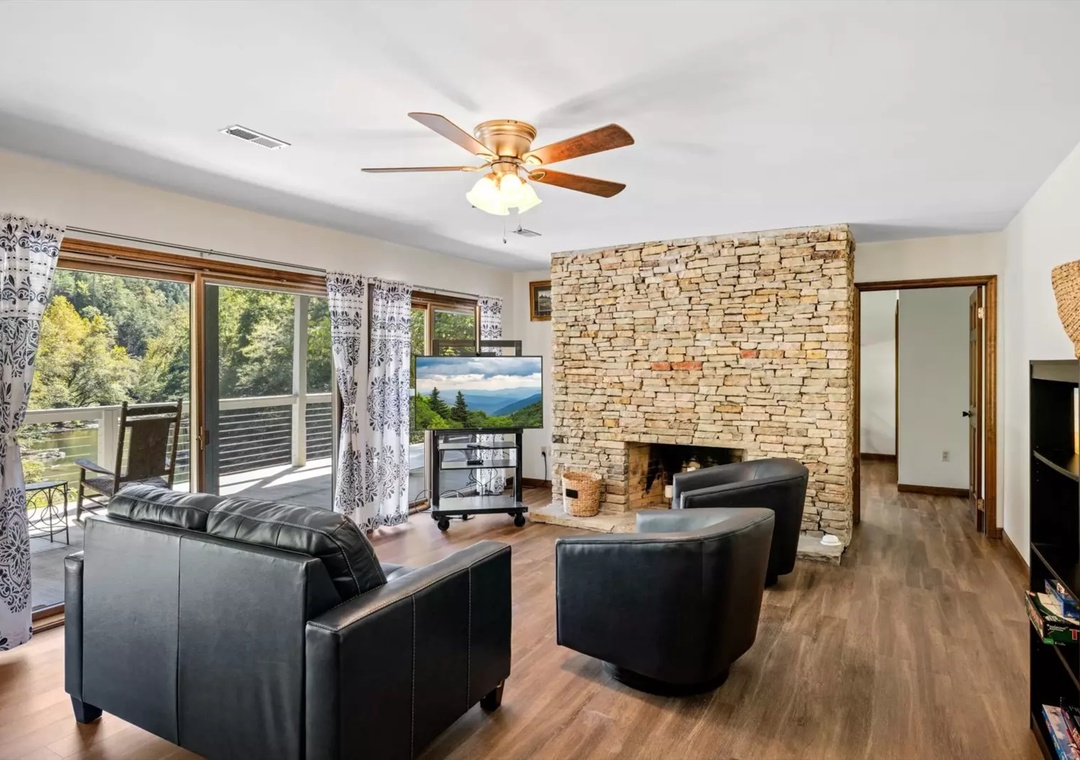 Unwind in your spacious living room featuring a stunning stone fireplace and comfortable leather seating, with serene forest views through sliding doors to your private deck.