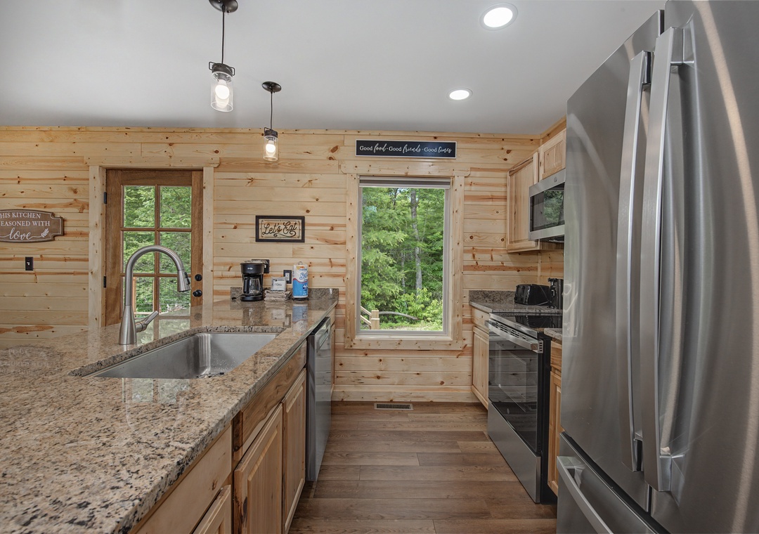 A warm, rustic kitchen awaits your culinary adventures with granite counters, modern appliances, and charming wood-paneled walls bathed in natural light from surrounding forest views.