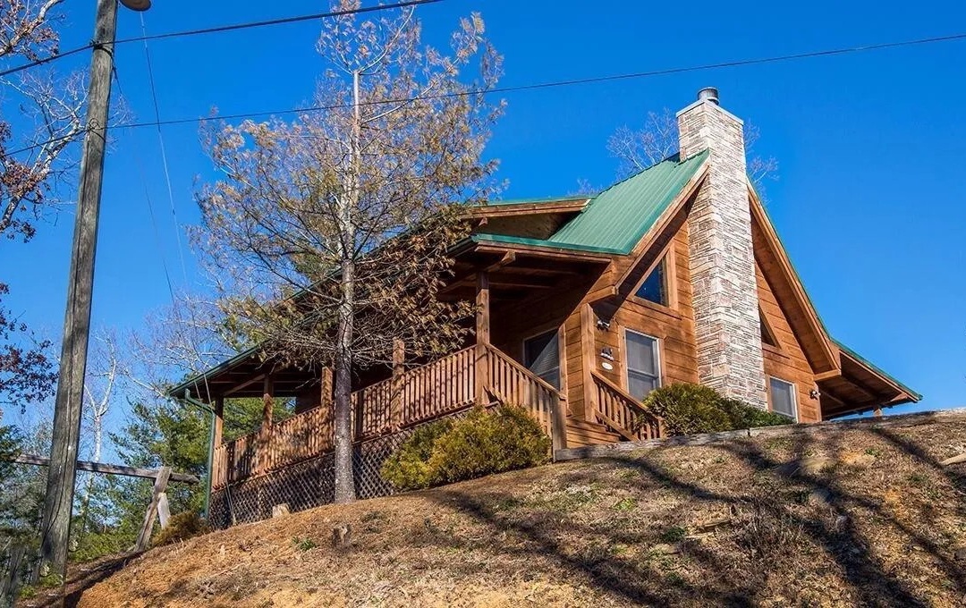 A rustic log cabin perched on a hillside, featuring natural wood construction, stone chimney, and wraparound deck surrounded by trees.