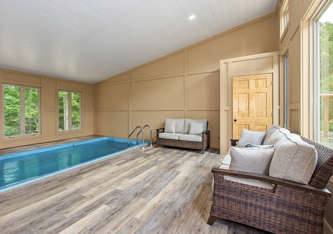 Step into your own private indoor pool oasis, where comfortable lounge seating invites you to unwind beside the crystal-clear water, surrounded by warm wood tones and lush garden views.
