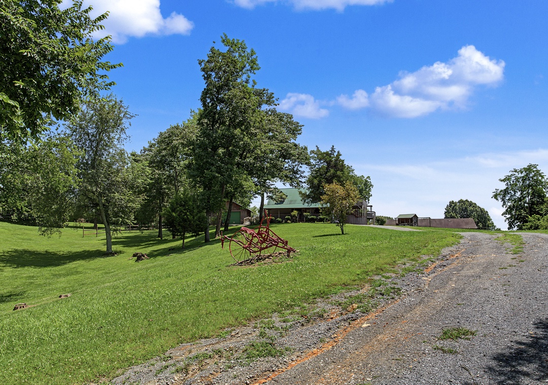 Spacious property surrounded by mature trees and open lawns, featuring a gravel driveway leading to the main building with additional outbuildings visible in the distance.