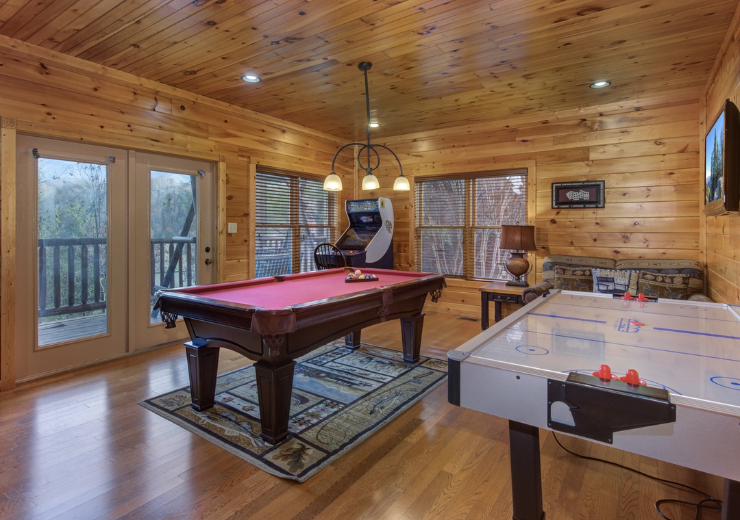 Step into your private game room, where friendly competition awaits with a full-sized billiard table, air hockey, and classic arcade entertainment—all wrapped in warm wood walls and natural light from the surrounding forest.