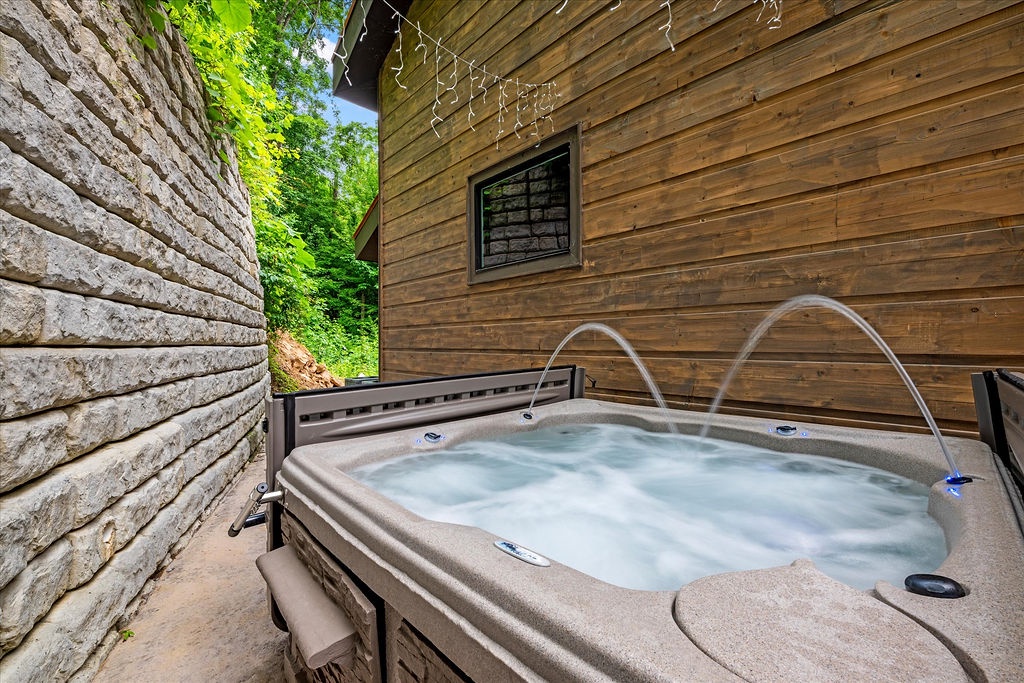 A secluded hot tub with water jets and fountain arcs, nestled between stone and natural wood siding on a private outdoor deck surrounded by lush greenery.