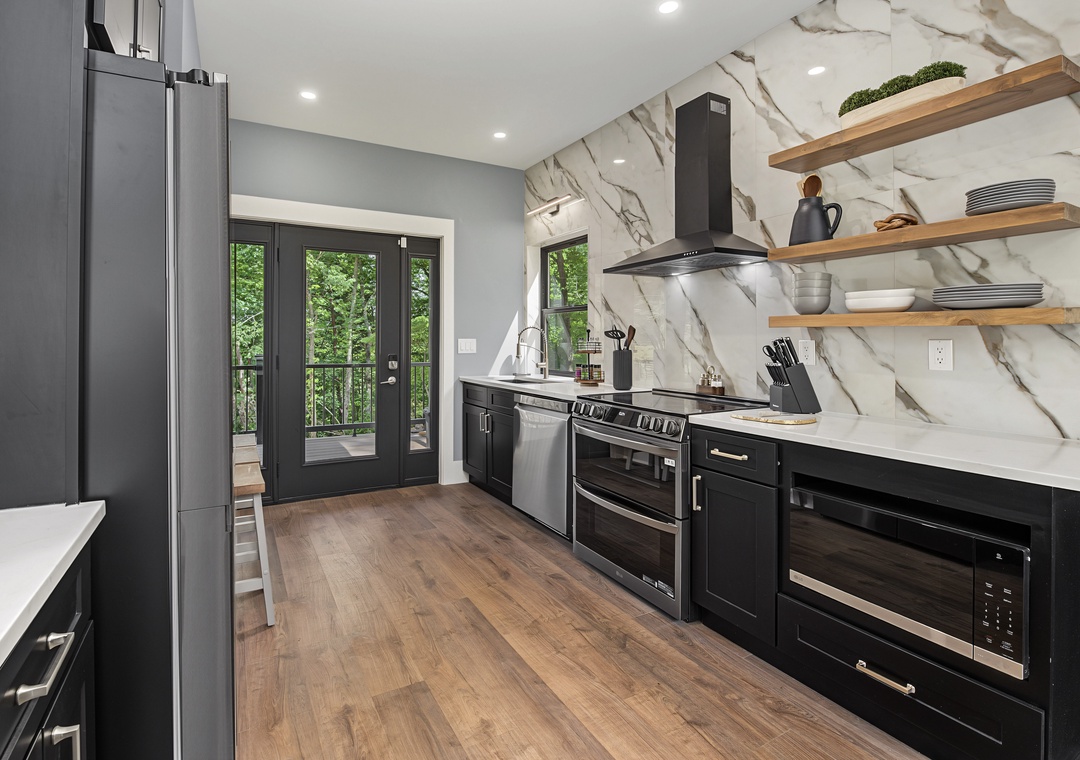 A sleek, modern kitchen awaits you with marble backsplash, wood shelving, stainless appliances, and French doors opening to a private balcony with leafy views.