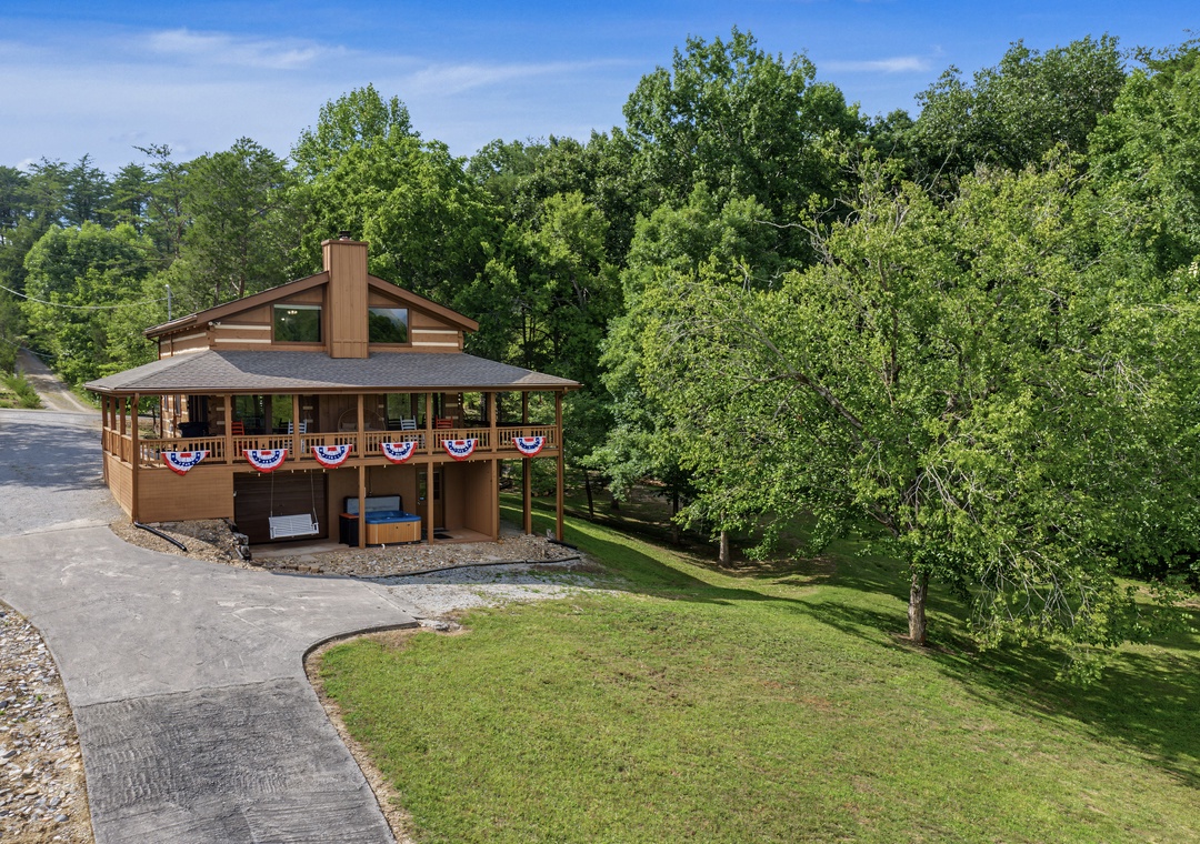 A welcoming cabin nestled among lush greenery, featuring a wraparound deck with festive bunting and ample parking, surrounded by mature trees and peaceful natural landscape.