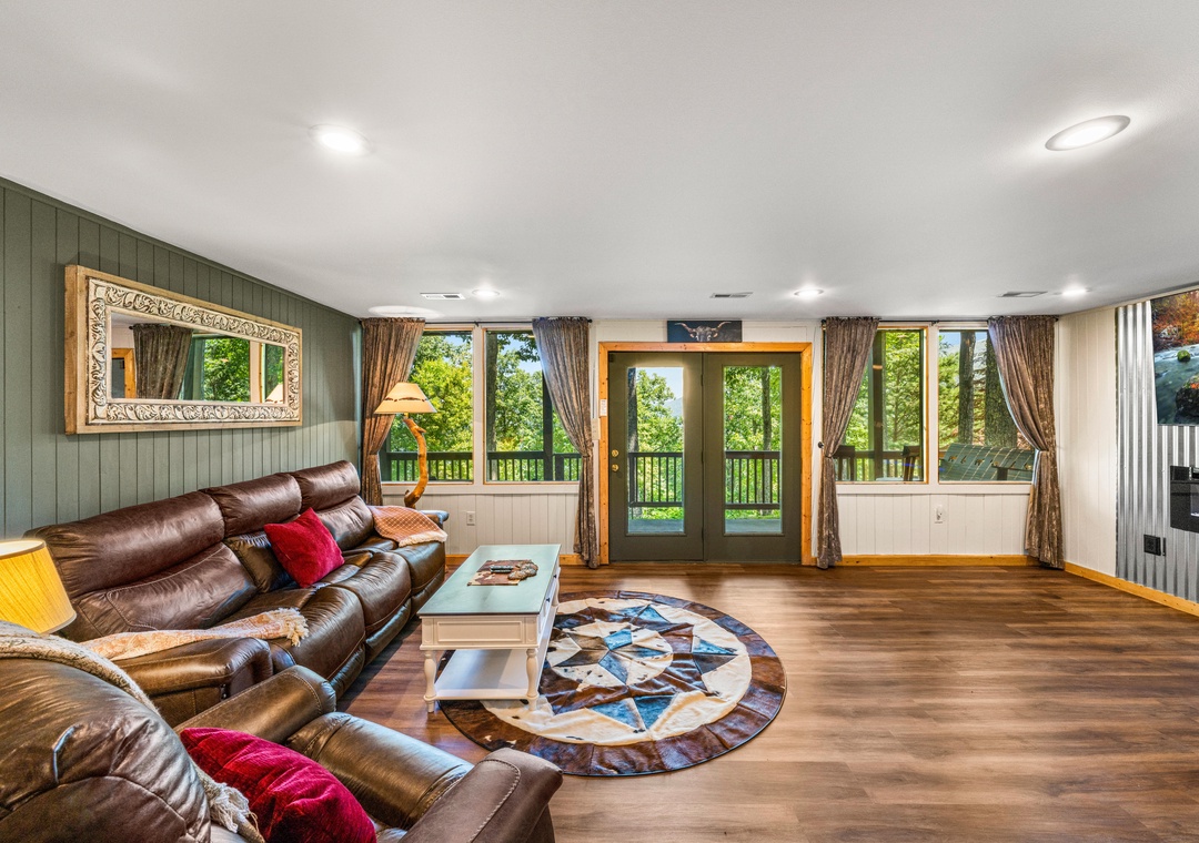 Step into your spacious living area with rich leather sofas, warm wood floors, and glass doors opening to a scenic balcony surrounded by lush greenery. Natural light fills the room.