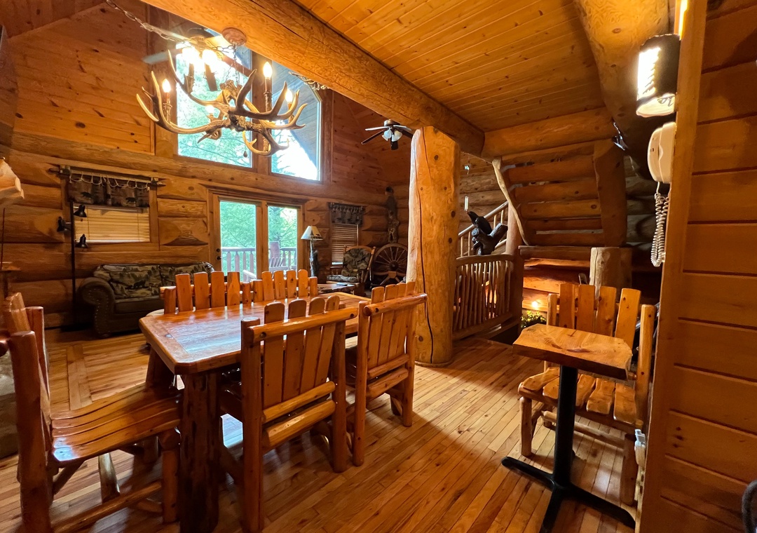 Rustic mountain retreat with hand-hewn log walls, soaring wood-beam ceilings, handcrafted dining table, and antler chandelier. Perfect for gathering and embracing authentic cabin living.