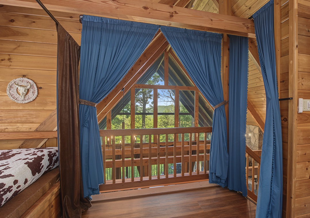 Step into your cozy loft bedroom where warm wood surrounds you and blue curtains frame stunning countryside views through the unique triangular window.