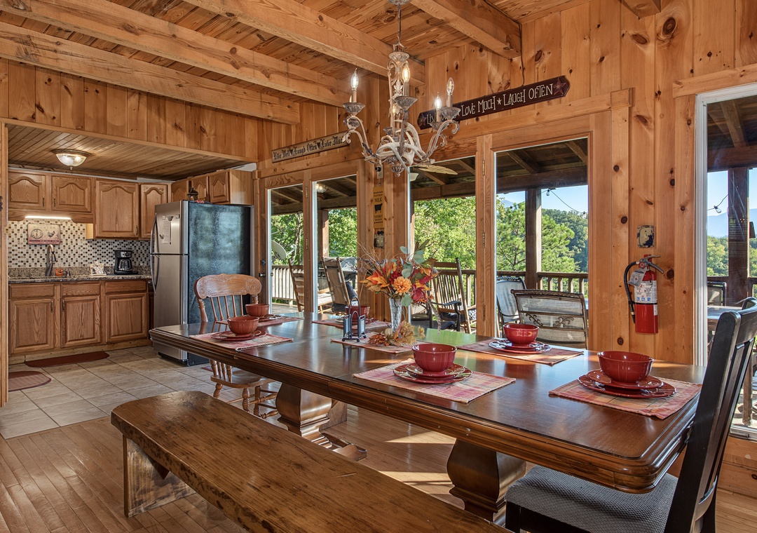 Gather around the rustic dining table under a charming chandelier, where warm wood beams and open views through glass doors invite you to linger over meals and shared stories in this cozy mountain retreat.
