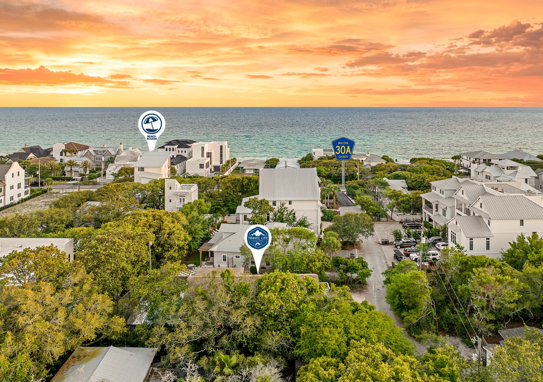 An aerial view captures a coastal neighborhood with beach homes nestled among lush greenery, just steps from the ocean at sunset.