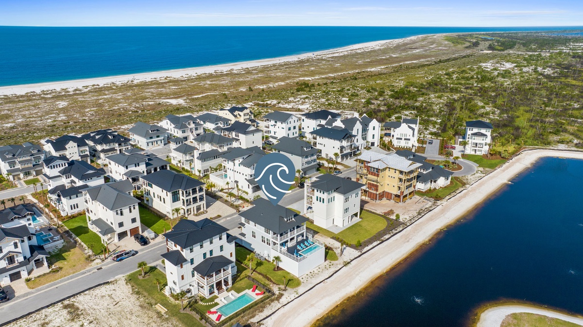 Aerial view of a coastal vacation rental community nestled between pristine beach and serene lake, surrounded by natural landscape and sandy shores.