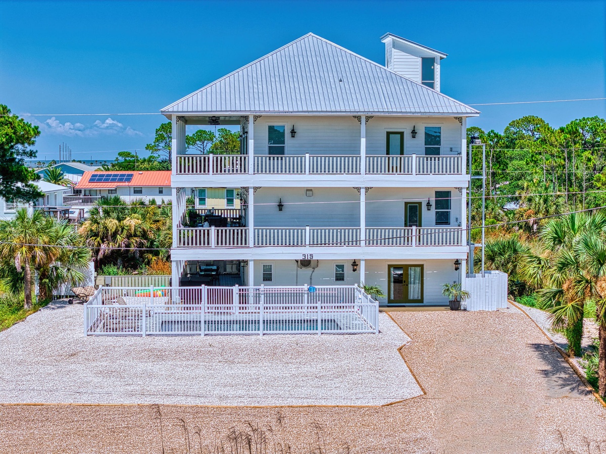 A striking three-story coastal property features wraparound balconies on each level, offering expansive outdoor spaces. Palm trees and lush greenery frame this beach-area home, complete with ample parking and coastal architecture.