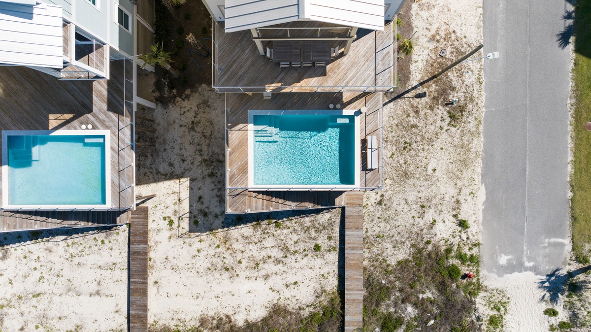 Aerial view of modern vacation rental property featuring private pools, wooden deck areas, and a beachfront location with sandy surroundings and adjacent street access.