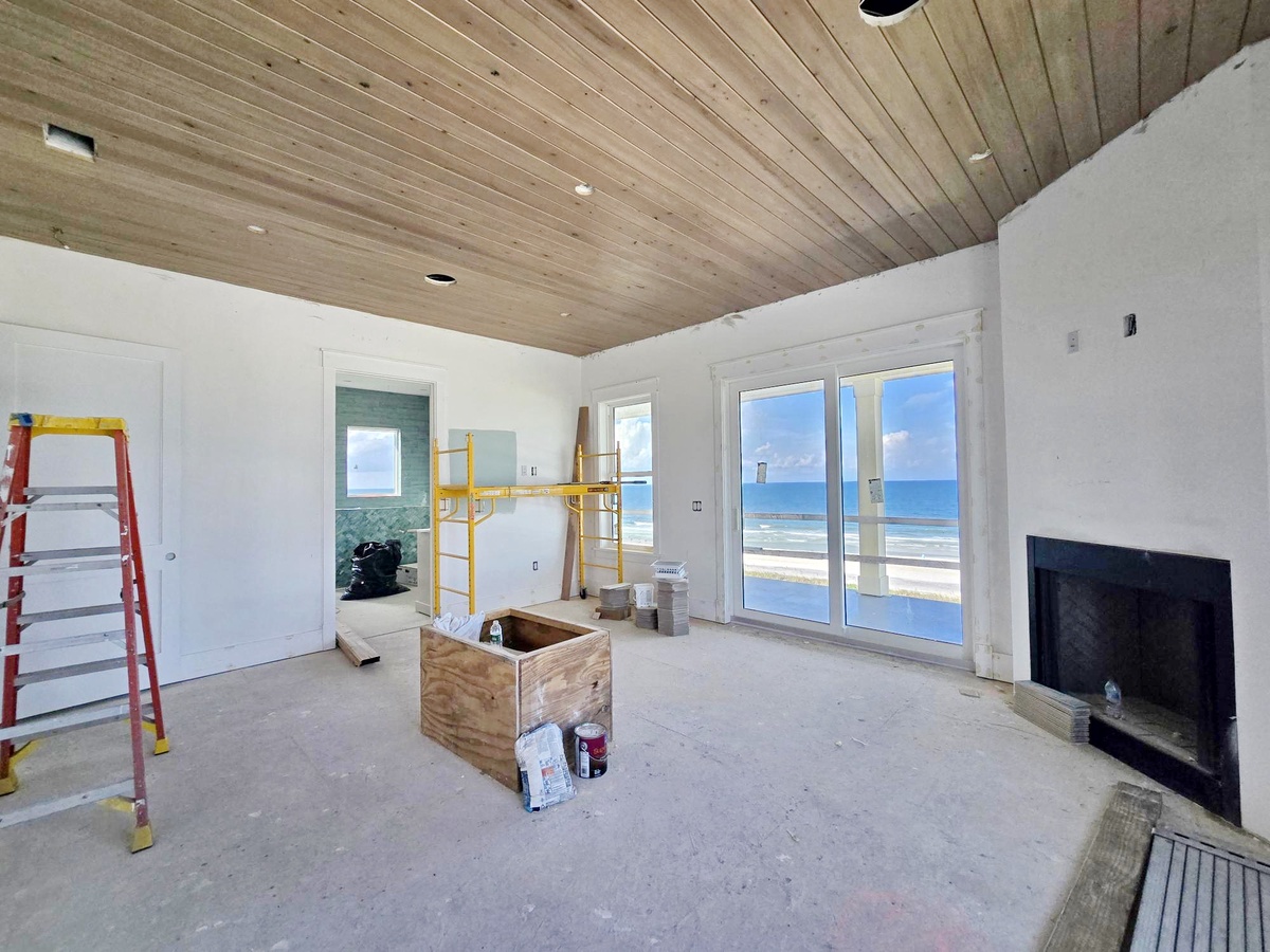 A spacious living room awaits finishing touches, featuring warm wood-planked ceilings and glass doors opening to stunning ocean views—imagine relaxing here as waves roll in.