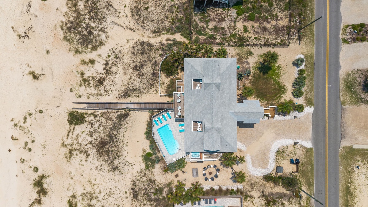 An aerial view captures a contemporary vacation property nestled in sandy coastal terrain, featuring a turquoise swimming pool and connecting boardwalk extending through natural dunes, with convenient road access nearby.