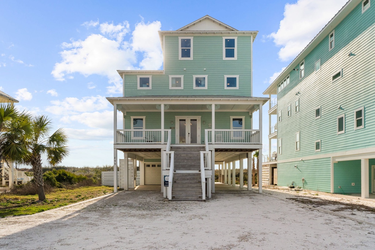 Charming coastal home with welcoming front porch and elevated design, perfectly situated among colorful beach houses in a peaceful neighborhood.