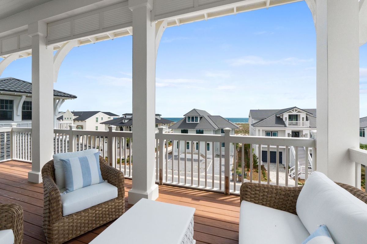 A scenic coastal neighborhood unfolds beyond the elevated balcony, with beach houses clustered near the shoreline and the ocean stretching toward the horizon.