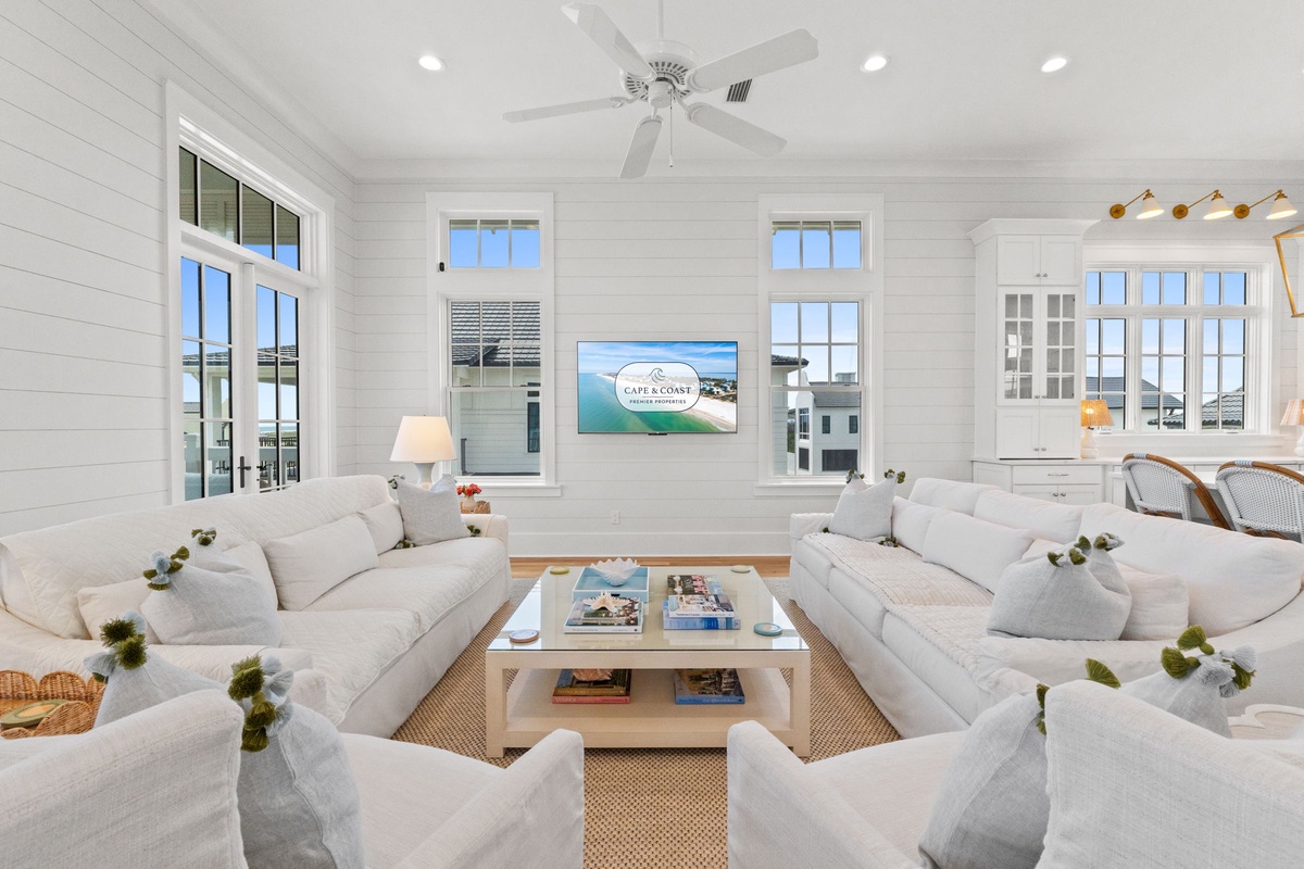 Sink into plush white sofas and unwind in this bright, coastal living room where natural light pours through tall windows and gentle breezes stir overhead.