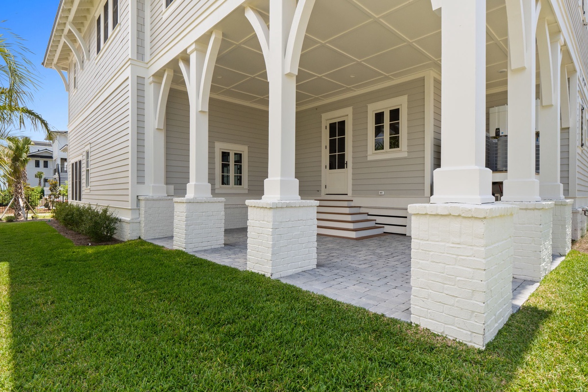This elegant coastal-style property features a grand wraparound porch with white brick columns and paver flooring, surrounded by manicured lawn and tropical landscaping under clear blue skies.