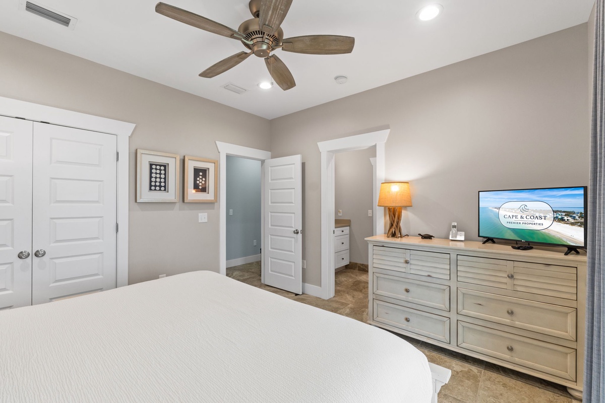 Your serene bedroom features a plush bed, coastal dresser with TV, warm lighting, and ceiling fan—perfect for unwinding after beach adventures.
