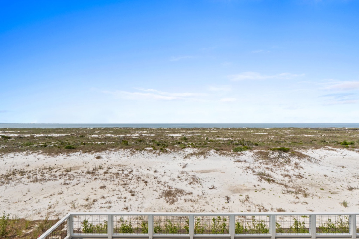 The property offers coastal surroundings with white sand dunes, native vegetation, and a clear horizon stretching across the Gulf, captured under a bright blue sky during daytime.