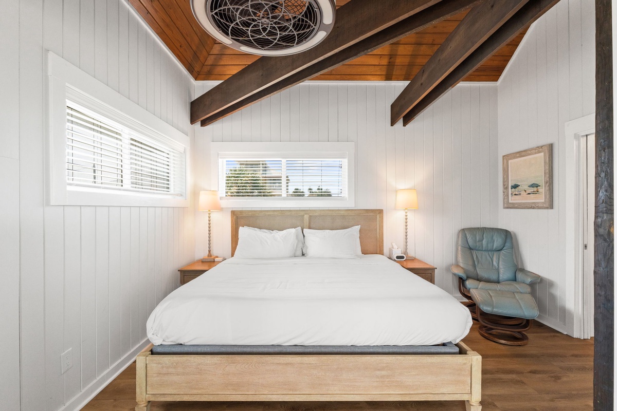 Unwind in your serene bedroom featuring vaulted wood-beamed ceilings, a plush king-sized bed with crisp white linens, and a cozy reading chair—your peaceful retreat awaits.