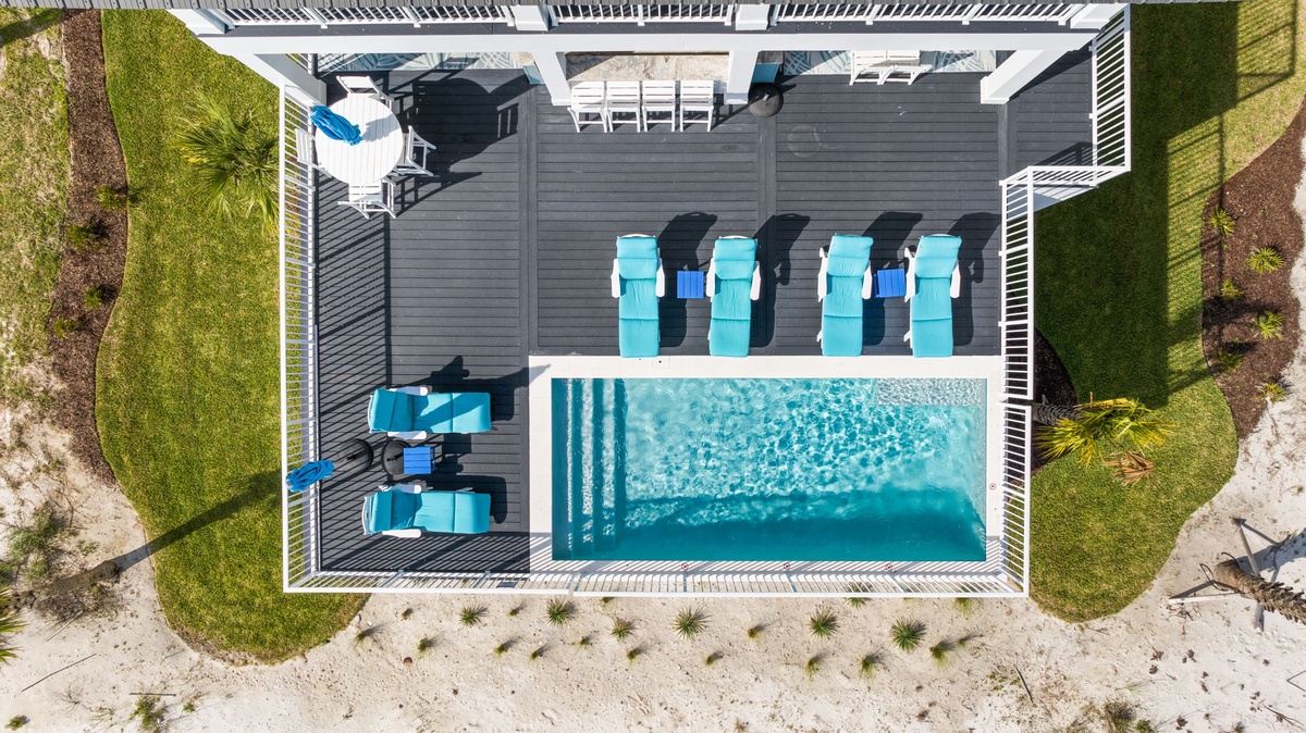 Aerial view of the property's pool deck featuring a pristine swimming pool with turquoise lounge chairs and white pergola, nestled between manicured lawns and sandy beach access.